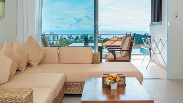 A modern living room with a beige sofa, a wooden coffee table with a flower vase, and a view of the ocean and pool area. - Hillside Seaview Residence (Karon)