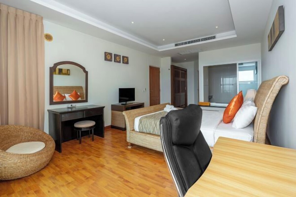 A hotel room with a wooden floor, a bed with orange pillows, a desk with a chair, a television, and a large mirror.