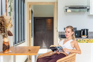Une personne est assise sur une chaise en osier, lisant un livre dans une pièce qui contient une table en bois, une fenêtre avec une plante et un coin cuisine équipé d'appareils électroménagers.