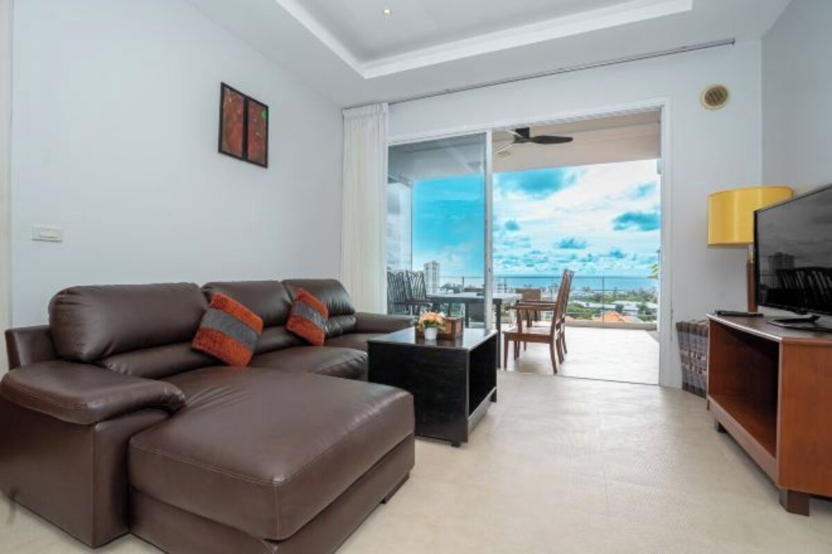 Een moderne woonkamer met een bruine leren bank, een zwarte salontafel en een tv-meubel. Er is een glazen schuifdeur naar een balkon met uitzicht op de stad.