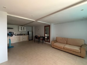 Uma sala de estar com sofá bege, área de jantar com mesa e cadeiras, cozinha com armários e eletrodomésticos, e um ventilador de teto.