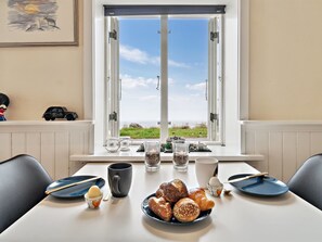 A dining table with pastries, a coffee mug, and a view of the sea through a window.