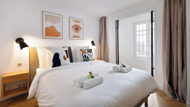 A hotel room with a large bed, two wall-mounted lamps, a window with curtains, and two framed artworks on the wall. - Appartement Le Marais - 1 Bedroom (Paris)