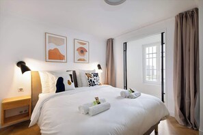 A hotel room with a large bed, two wall-mounted lamps, a window with curtains, and two framed artworks on the wall. - Appartement Le Marais - 1 Bedroom (Paris)