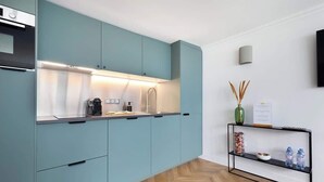 A modern kitchen with teal cabinets, a stainless steel countertop, and a black metal shelf with decorative items. - Appartement Le Marais - 1 Bedroom (Paris)