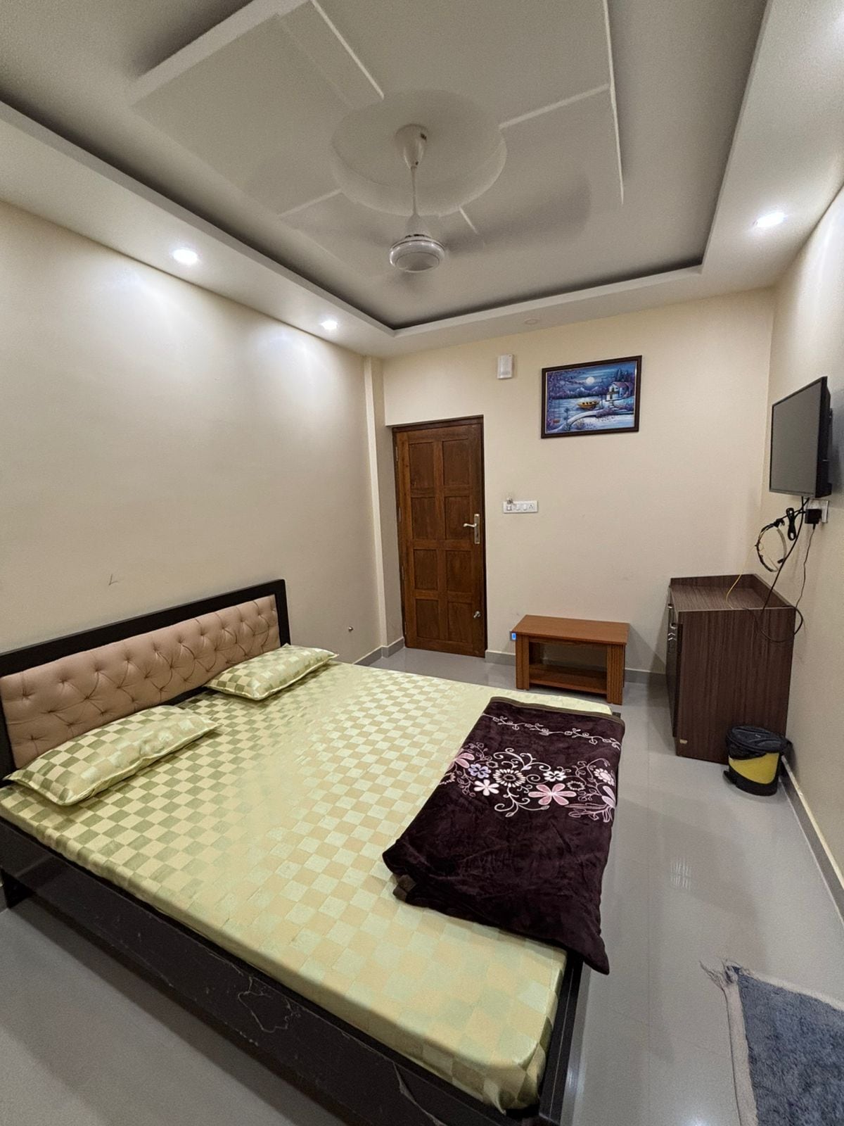 A bedroom with a bed, a ceiling fan, a wooden door, a small table, a TV, and a bag.