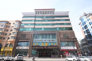 Un edificio de varios pisos con una amplia entrada de vidrio, varios rótulos comerciales y algunos automóviles estacionados frente a él.