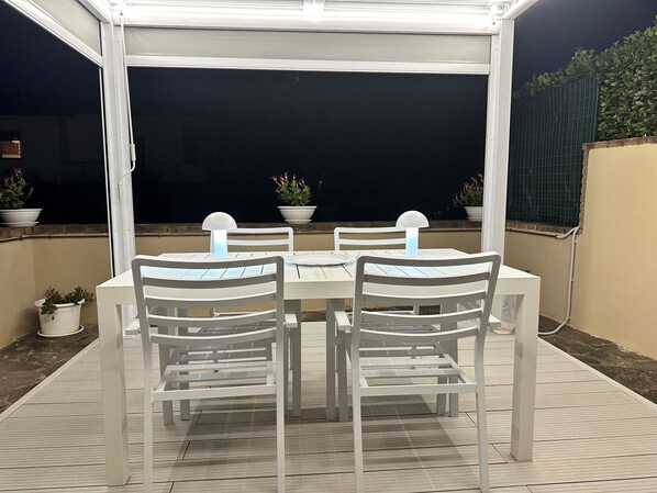 Un patio al aire libre con una mesa y cuatro sillas, rodeado de plantas en macetas y una zona cubierta.