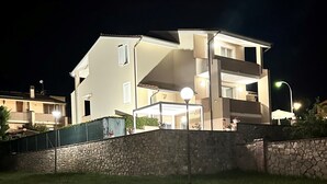 A multi-story residential building with a stone wall in front, a balcony on the second floor, and a lit entrance.