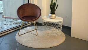 A modern room with a large sliding glass door, a round white rug, a brown chair, a small white table with a plant, and a marble-like wall art piece.