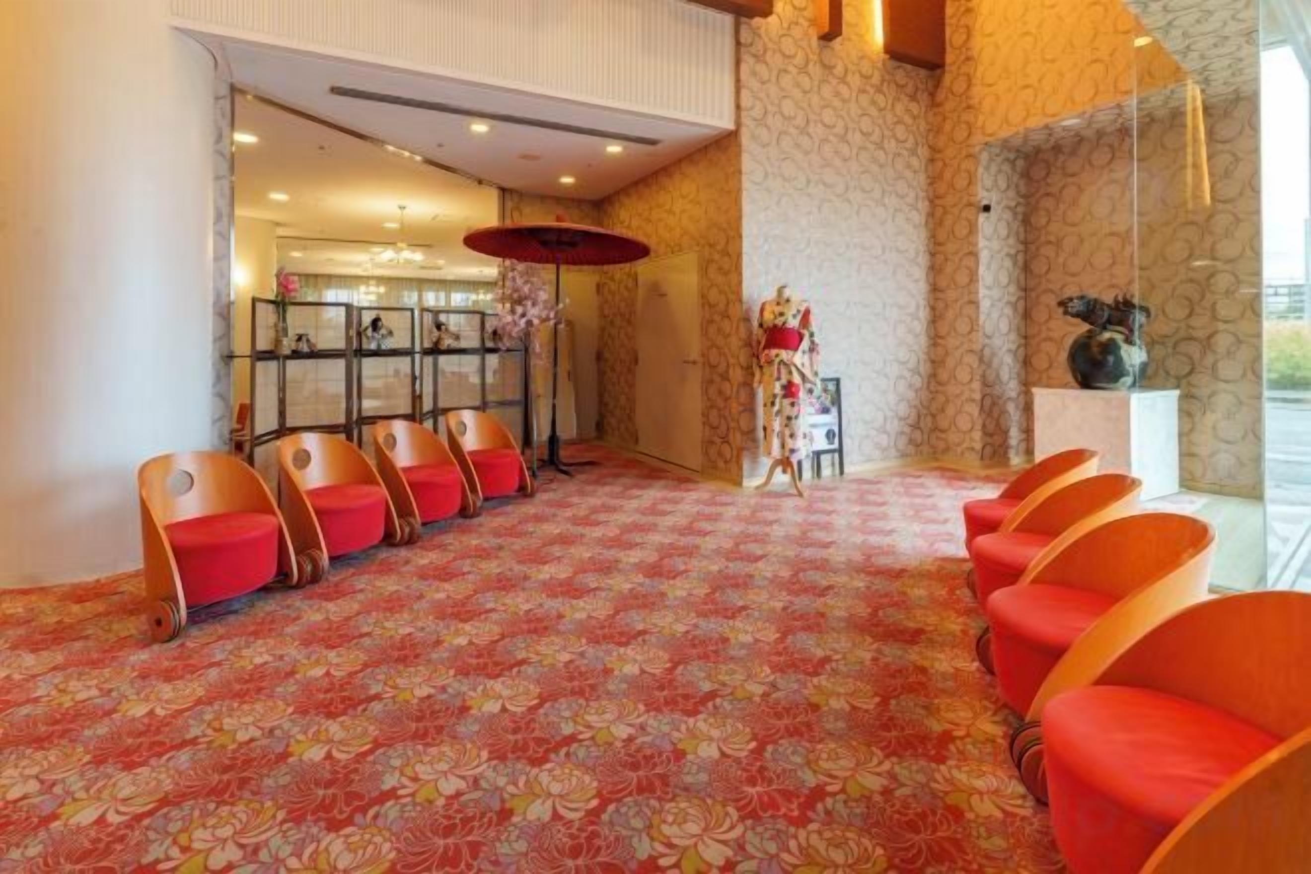 Un hall de hotel con alfombra estampada, filas de sillas rojas, una mesa de recepción y un jarrón decorativo.