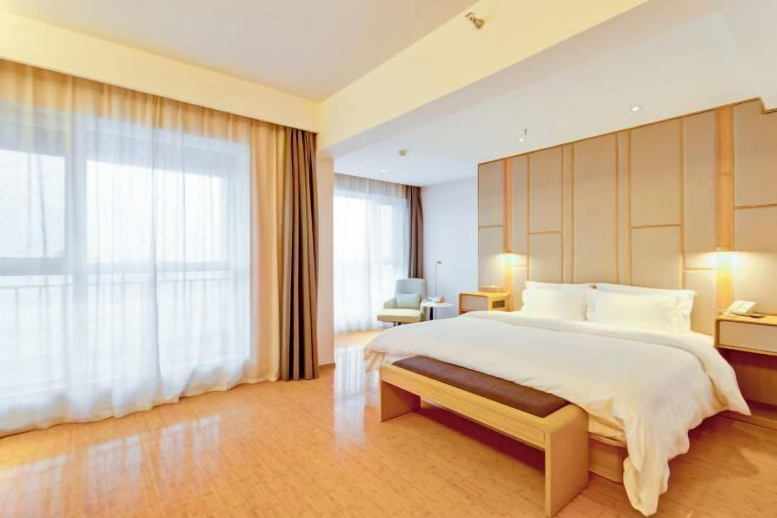 A modern hotel room with a large bed, a bedside table, a chair, and a large window with curtains.