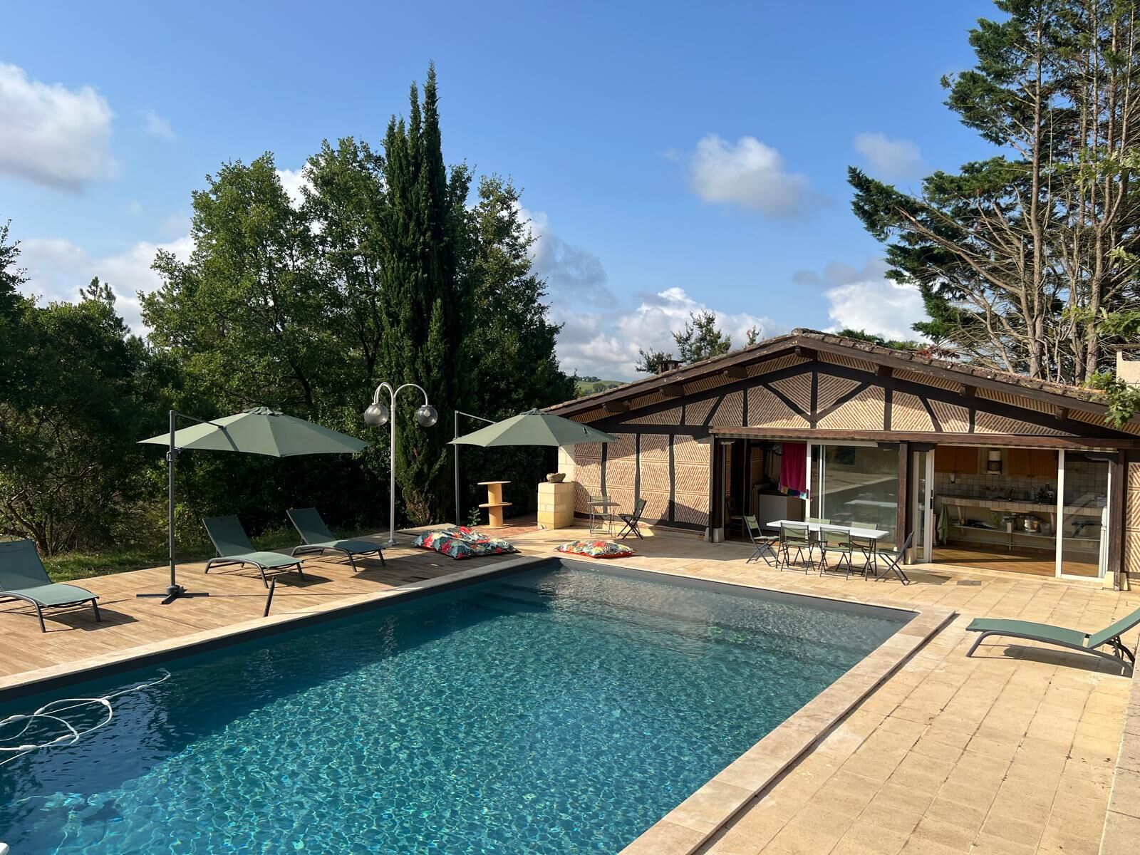 A swimming pool with lounge chairs and umbrellas, a covered patio area, and a view of the surrounding trees.