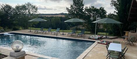 Une piscine rectangulaire, agrémentée de chaises longues et de parasols, entourée d’une terrasse en bois et de murs en pierre.