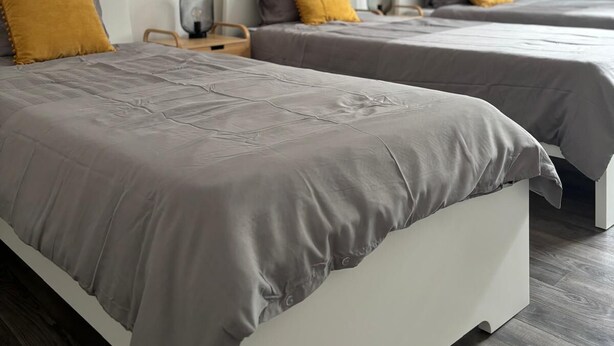 Une chambre moderne avec deux lits, chacun recouvert d’une couverture grise et agrémenté d’un coussin de couleur moutarde. Les murs de la pièce sont blancs, une carte du monde est accrochée au mur et le sol est en bois.