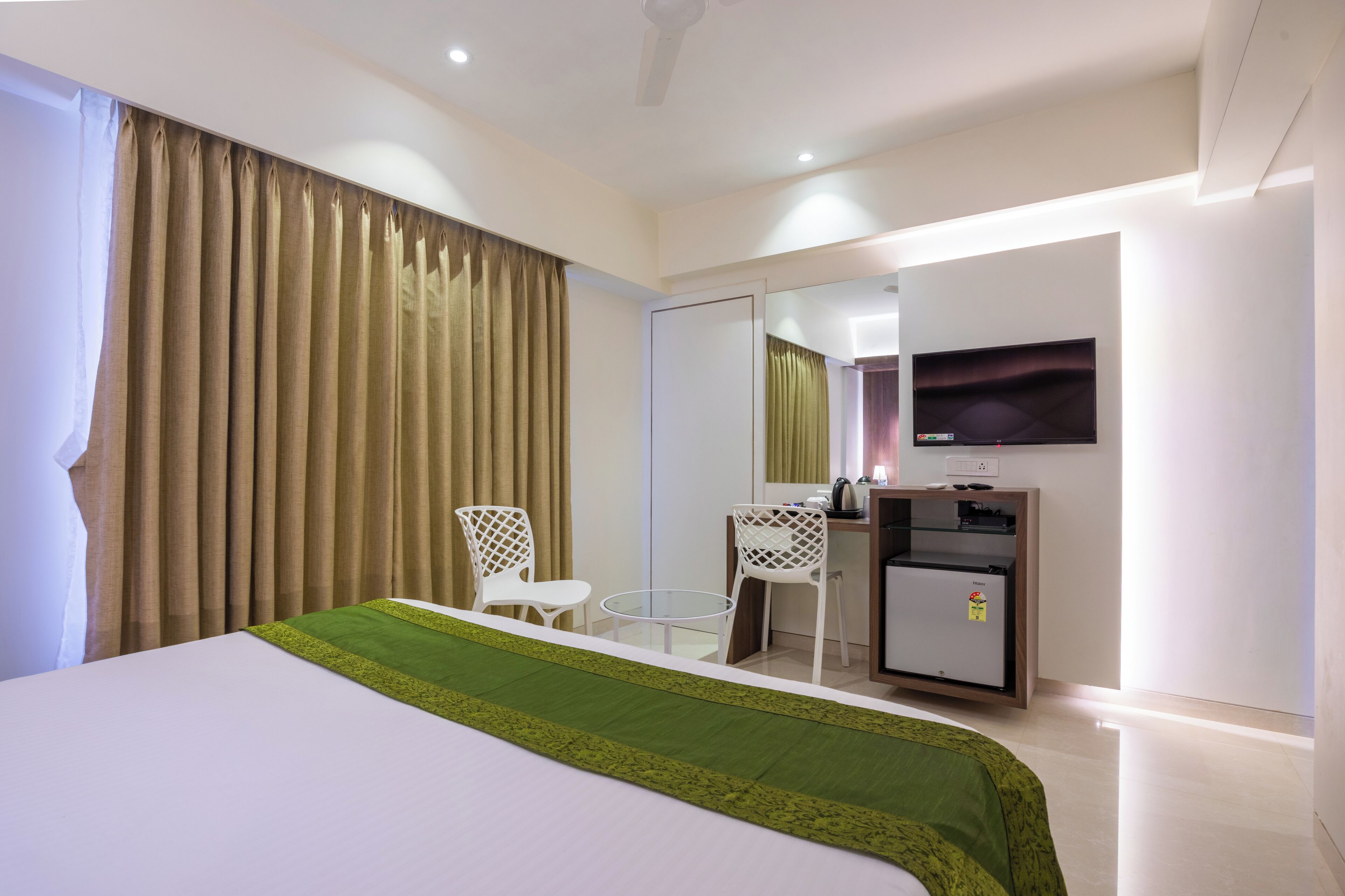 Habitación de hotel con cama, escritorio, silla, refrigerador pequeño y televisor en la pared.
