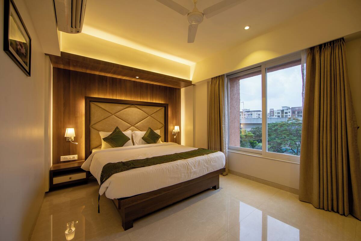 A hotel room with a large bed, a wooden headboard, two bedside lamps, a ceiling fan, and a large window with curtains.
