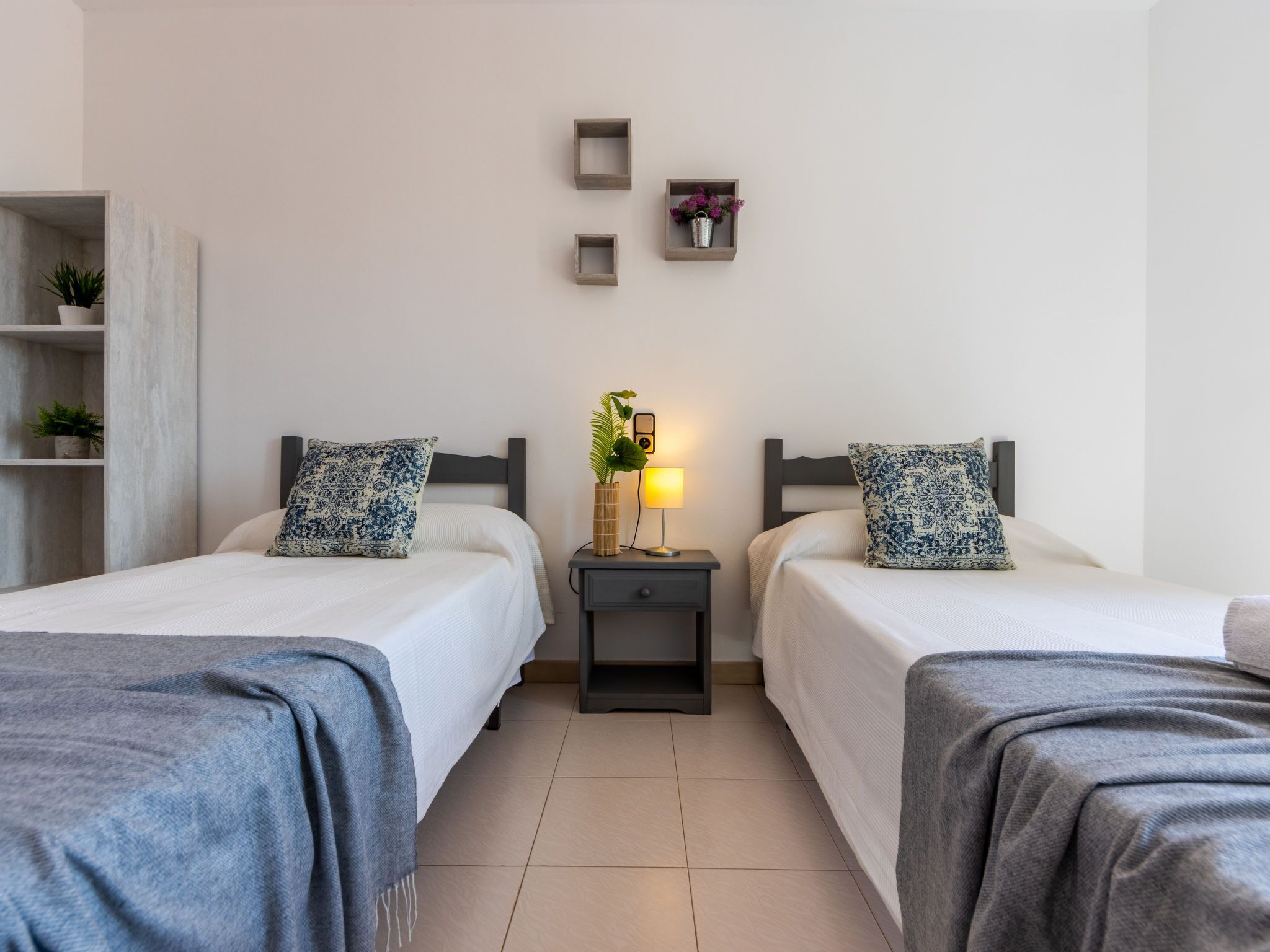 Habitación de hotel con dos camas individuales, una mesita de noche con lámpara, un pequeño estante con una planta en maceta y una repisa en la pared con un florero.