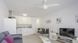 A modern living room with a grey sofa, a glass coffee table, and a flat-screen TV. There is a kitchen area with white cabinets and a white ceiling fan. - Parque Don Jose Poolside Retreat by Interhome (Costa del Silencio)