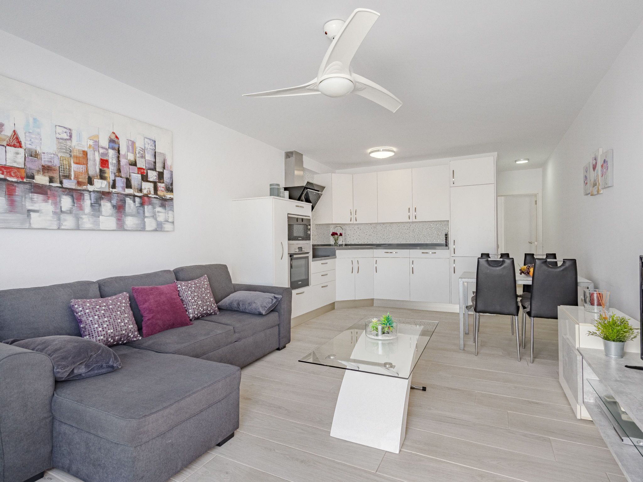 A modern living room with a grey sofa, a glass coffee table, and a ceiling fan.