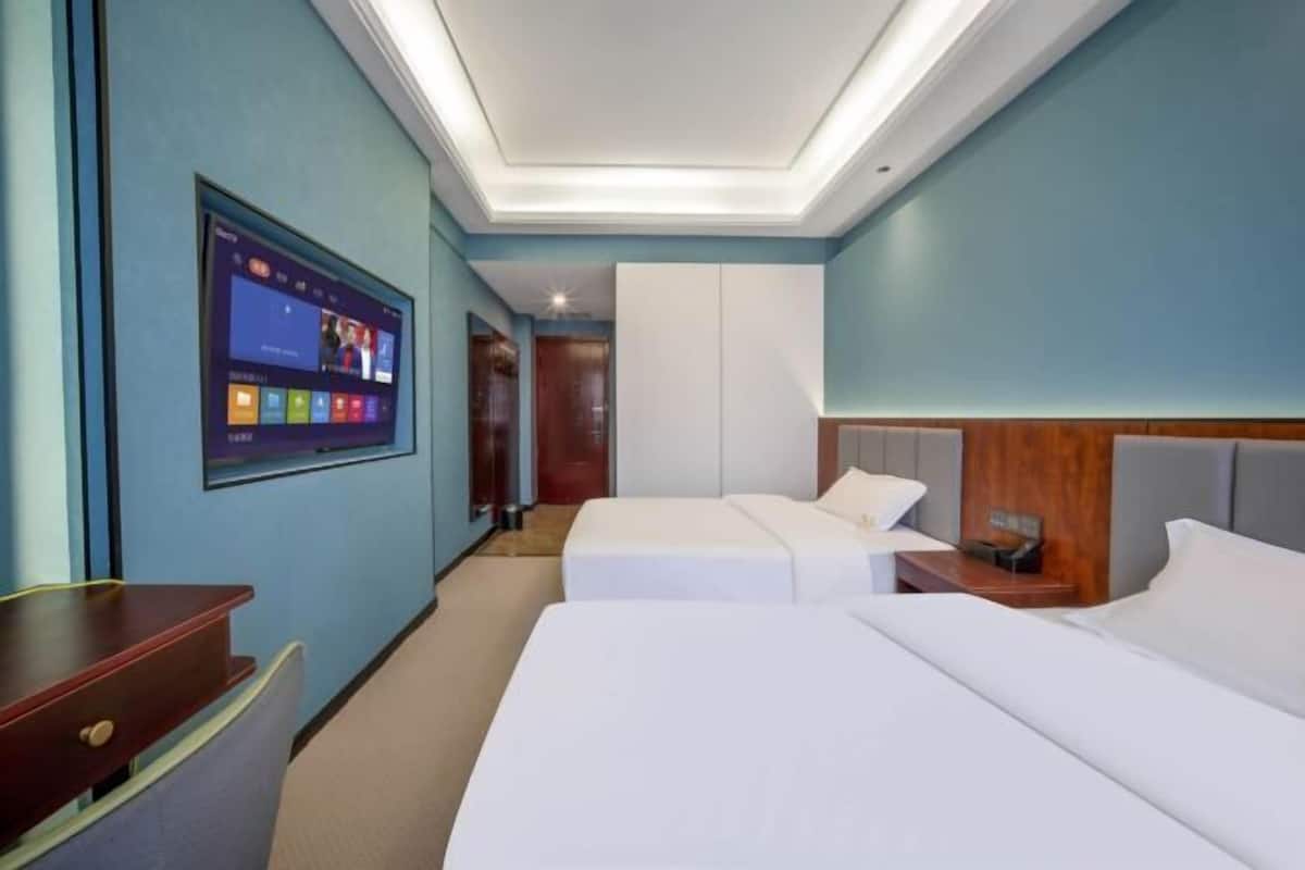 A hotel room with two beds, a desk, a TV mounted on the wall, and a ceiling light.