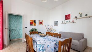 A dining area with a table covered in a floral tablecloth, surrounded by wooden chairs. There is a grey sofa against the wall and a door with a teal door frame. - 4061 Casa Wilma by Barbarhouse (Porto Cesareo)