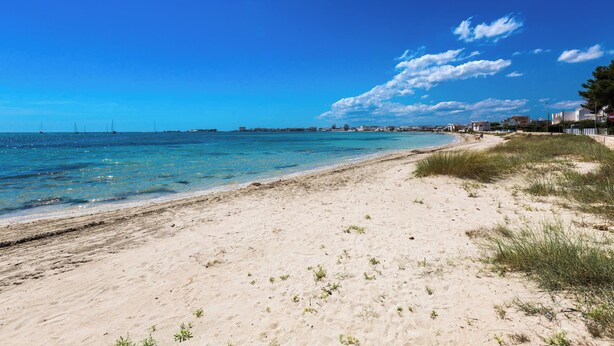 A beach with clear blue water, sandy shore, and buildings in the background. - 4061 Casa Wilma by Barbarhouse (Porto Cesareo)