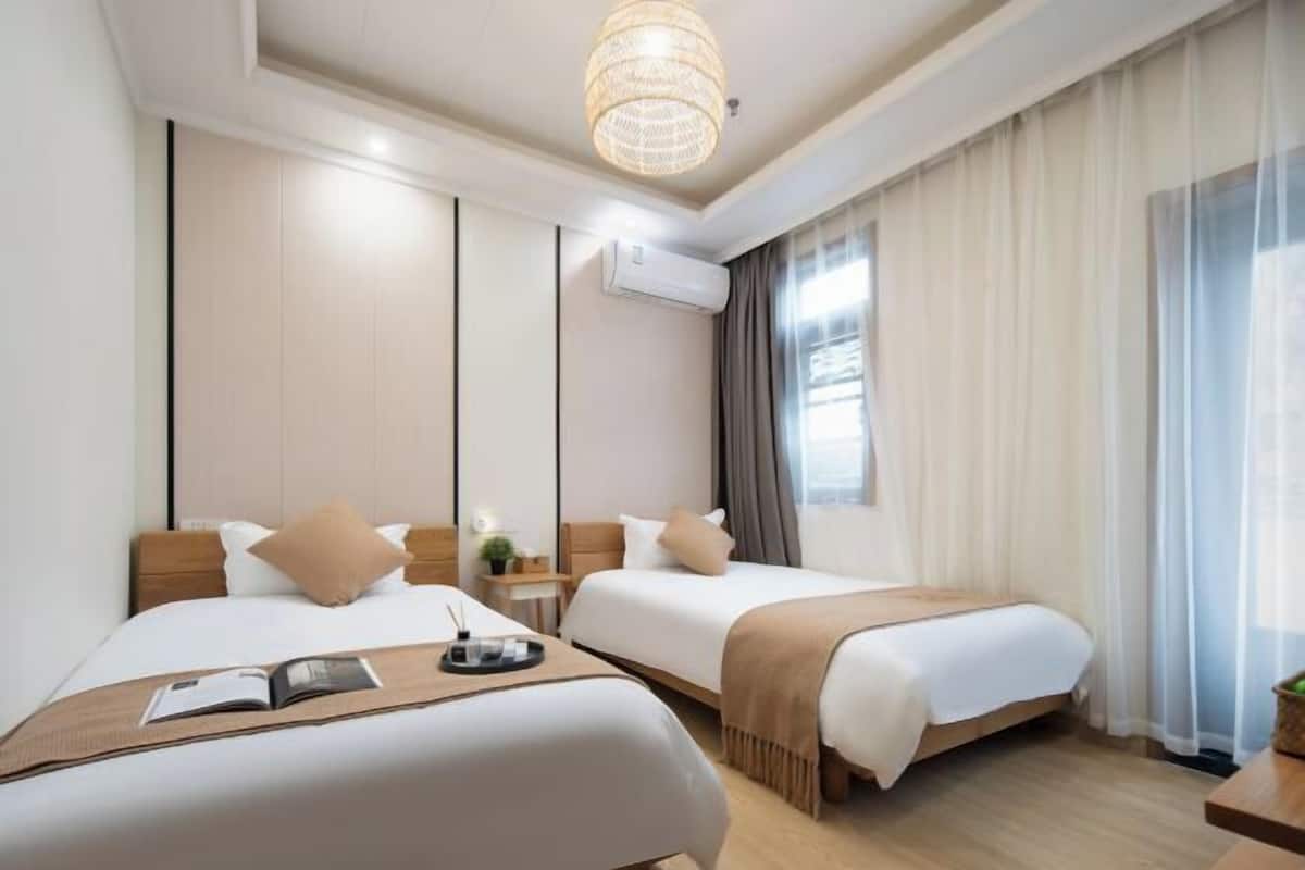 Una habitación de hotel moderna con dos camas, un ventanal con cortinas translúcidas, una lámpara colgante y un aire acondicionado de pared.