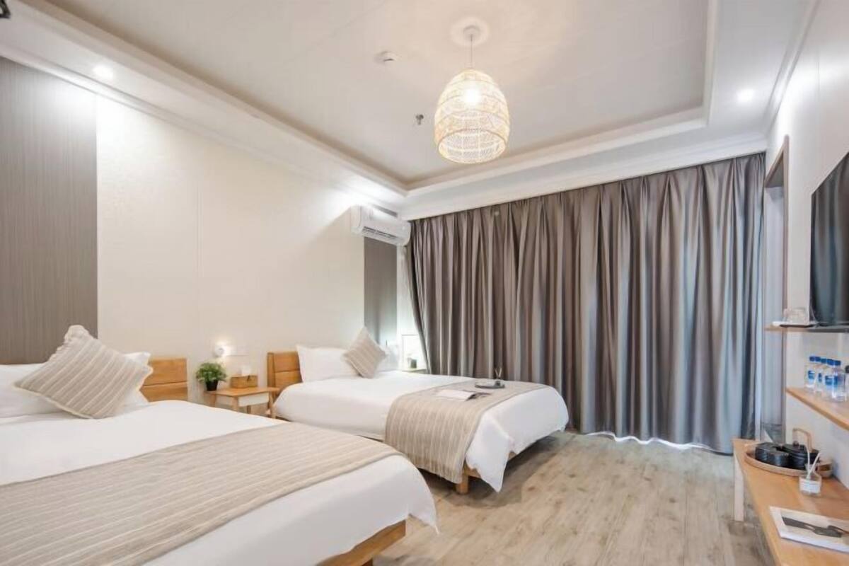 Habitación de hotel con dos camas, un escritorio, una silla, una lámpara colgante y un ventanal con cortinas.