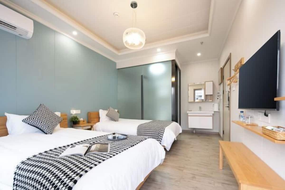 Habitación de hotel con dos camas, un banco de madera, un televisor, un escritorio y un baño visible a través de una puerta abierta.
