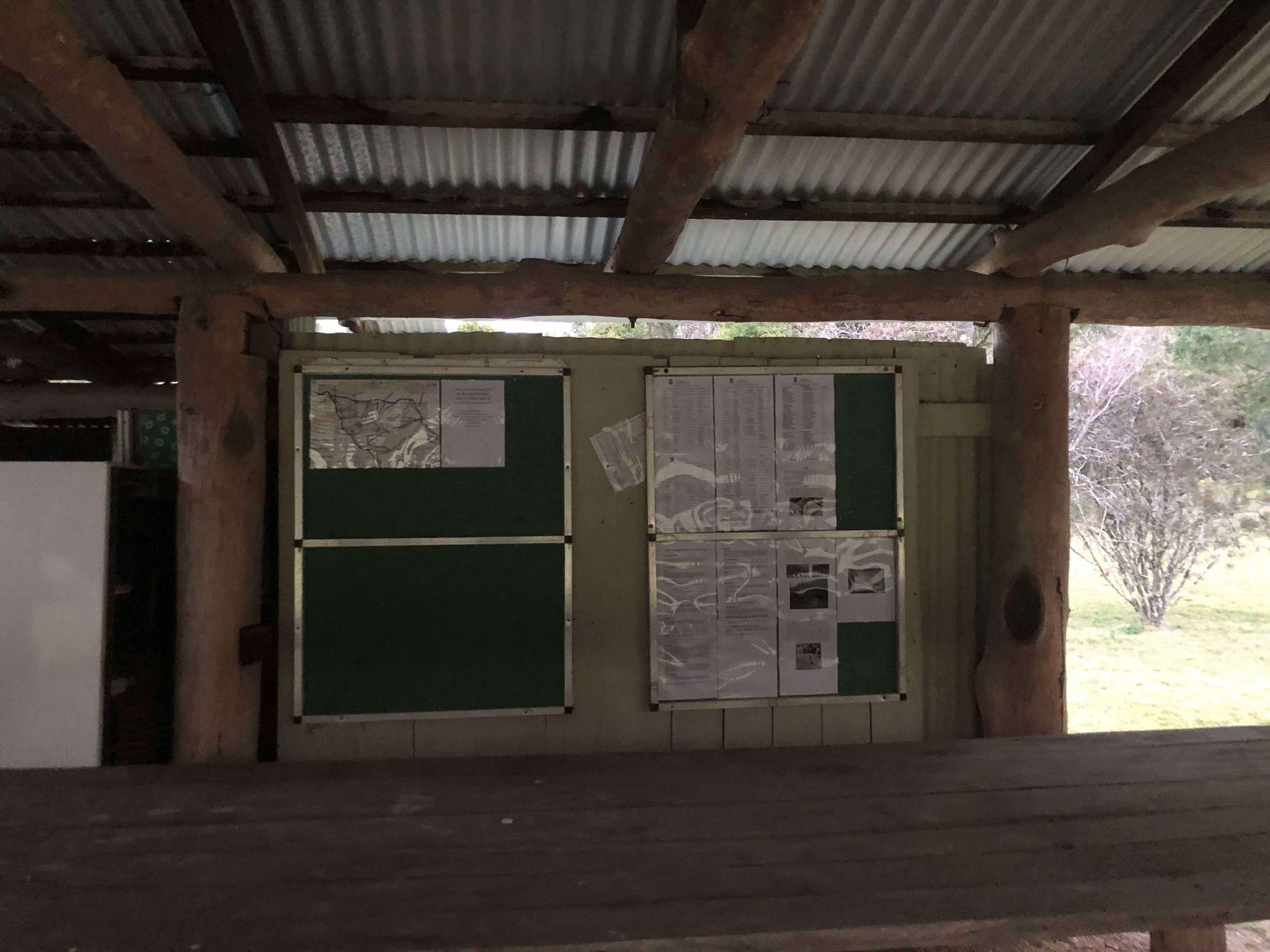 Un espace extérieur abrité, doté d’un banc en bois, d’un panneau d’affichage où sont accrochés des documents et d’un toit en métal.