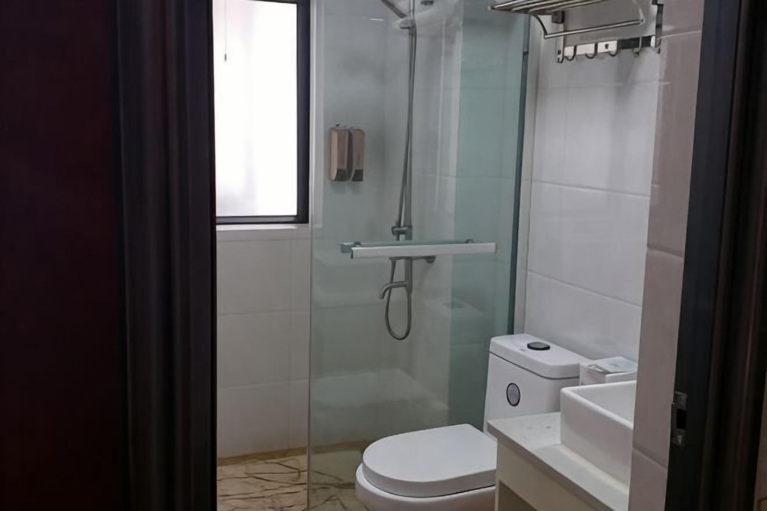 Um chuveiro com divisória de vidro, vaso sanitário, pia, dispensador de toalhas e janela.