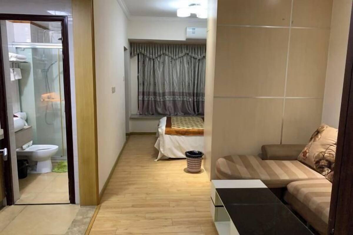 A small, compact living space with a wooden floor, a bed with a striped blanket, a sofa with patterned cushions, a small table, and a window with curtains.