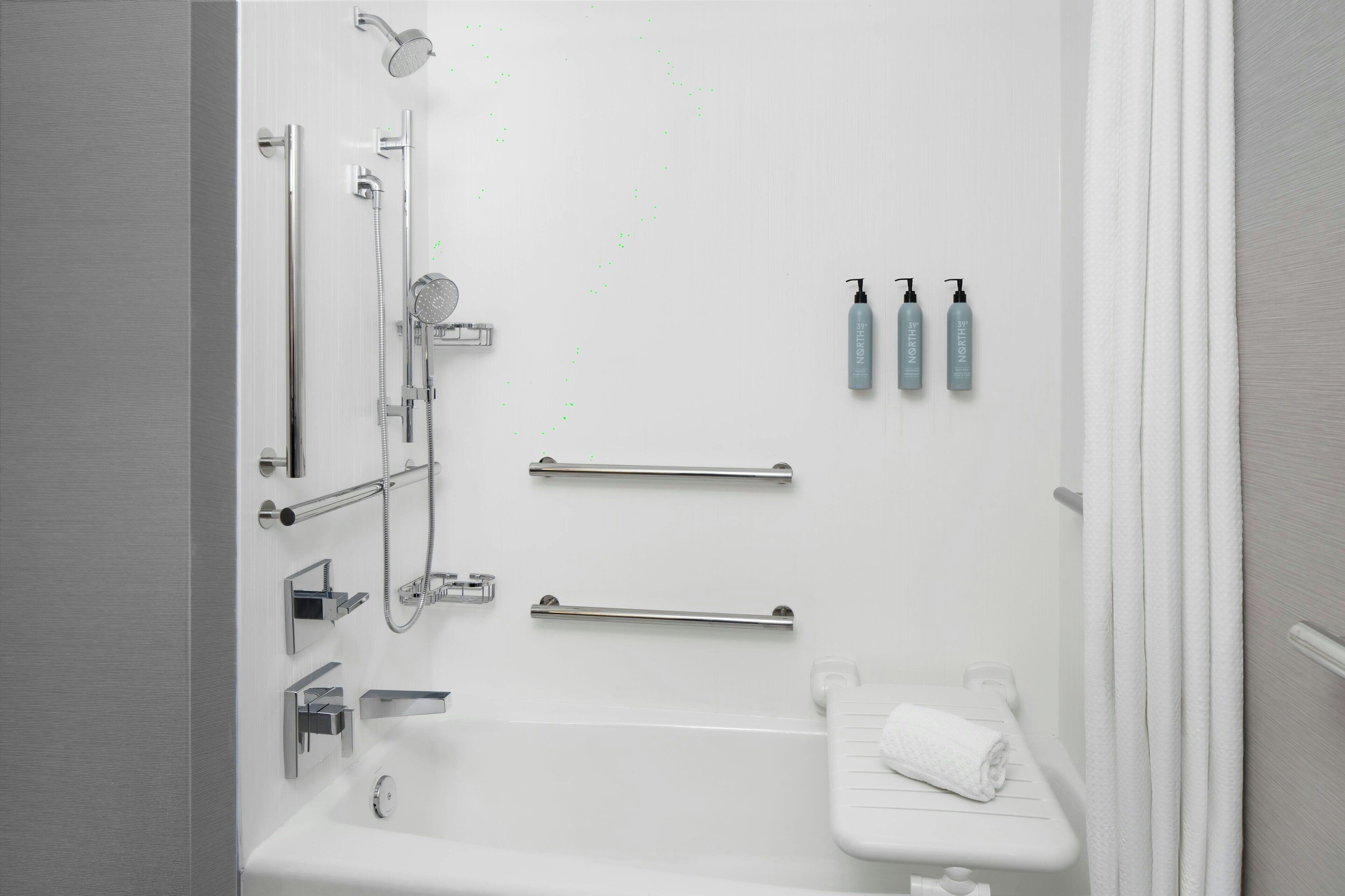 Een moderne badkamer met een bad, douche en toilet. Er zijn twee handdoekhouders, een handdroger en drie flessen handzeep aan de muur.