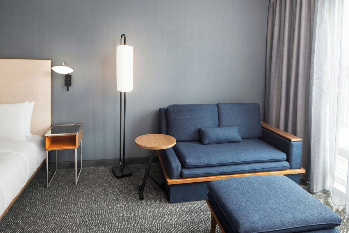 Una habitación de hotel con una cama, un sofá azul, una pequeña mesa de madera, una lámpara de pie y un ventanal con cortinas.