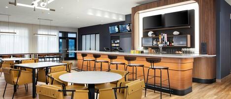 A modern restaurant with round tables and yellow chairs, a bar area with a large TV, and a wooden floor.