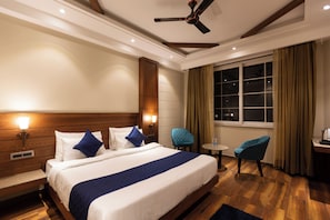 Habitación de hotel con cama, lámparas de noche, ventilador de techo, ventana con cortinas, una mesita y una silla.