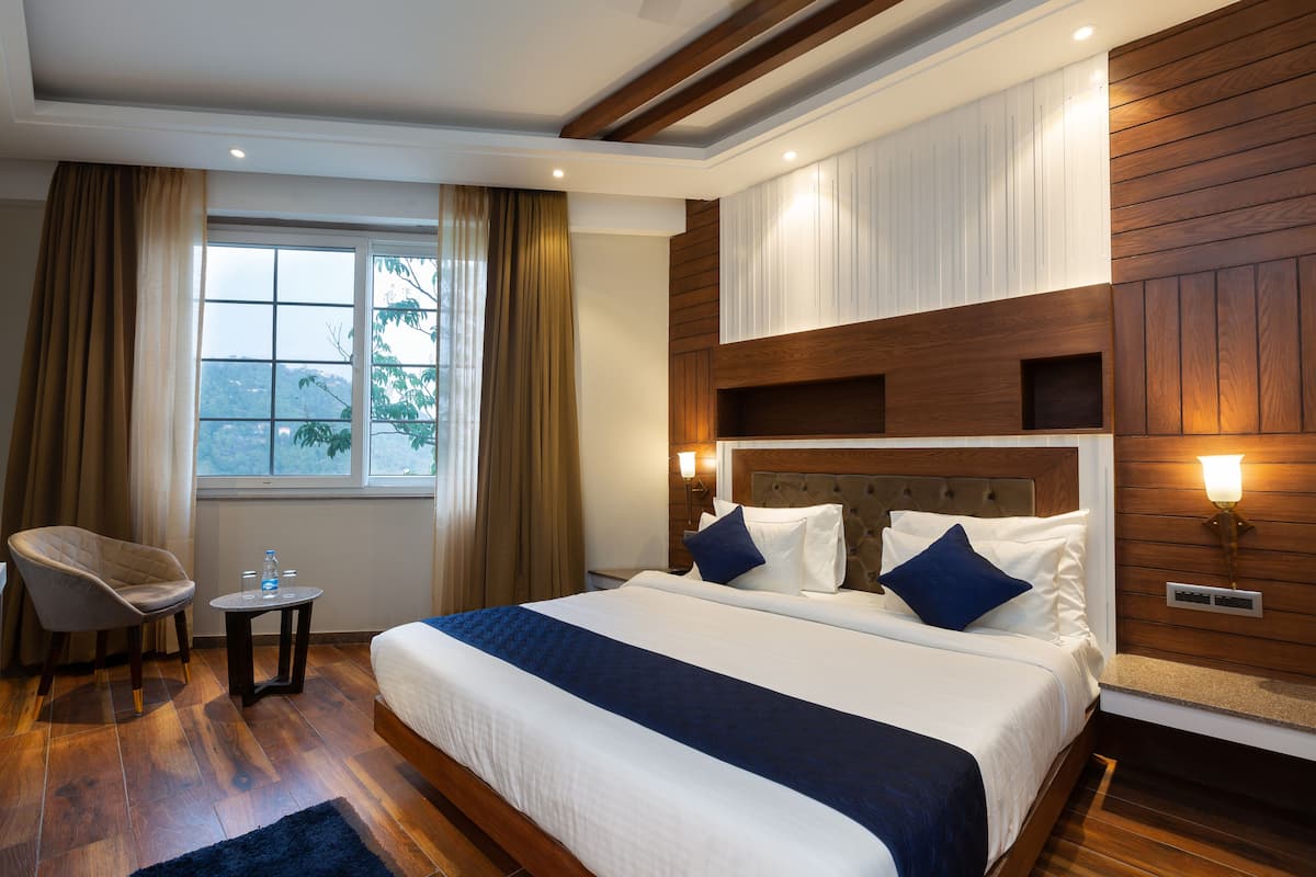 A hotel room with a large bed, a chair, a small table with water bottles, and a view of the outdoors through a window.