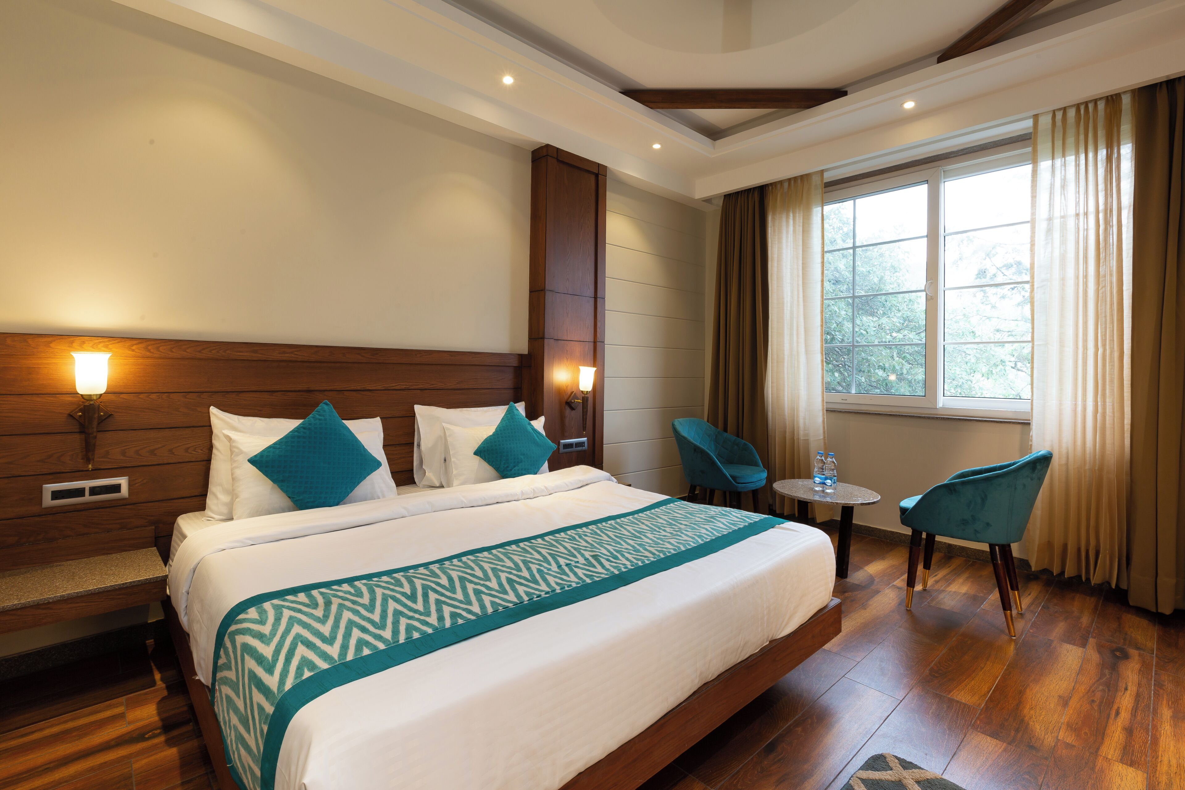 A hotel room with a large bed, two chairs, a small table, and a window with curtains.