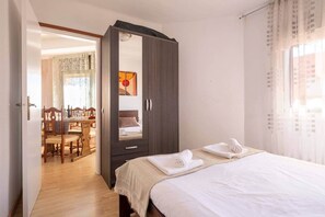 Une chambre dotée d’une grande armoire, d’un lit avec du linge de lit blanc, et d’un coin repas visible à travers une porte ouverte.