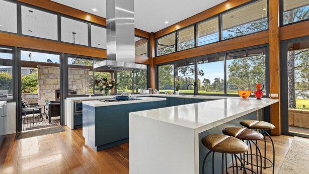 A modern kitchen with a large island, a dining area with a stone fireplace, and large windows offering a view of the outdoors. - HUNTER VILLA - THE VINTAGE (POKOLBIN)