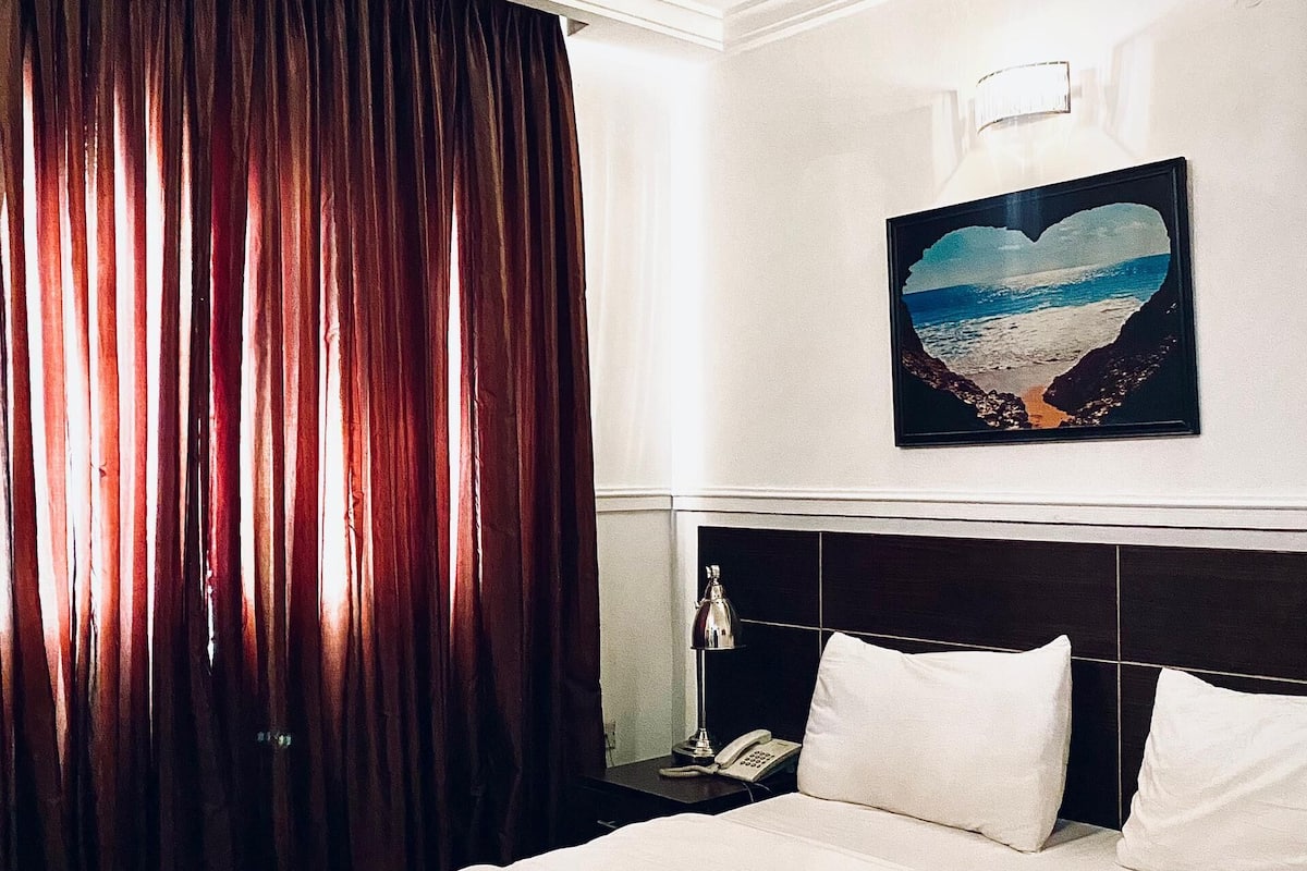 Habitación de hotel con cama, mesita de noche, teléfono, un cuadro en la pared y ventana con cortinas.