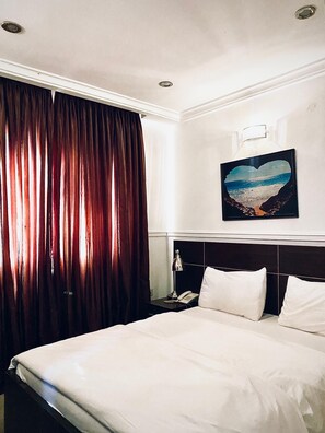 Una camera d'albergo con un letto, un comodino, un telefono, un quadro appeso al muro e una finestra con tende.