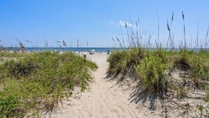 Un sentier de sable qui longe la plage et mène à l’océan, avec des parasols et des chaises de plage en arrière-plan.