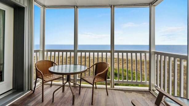 Un espace extérieur abrité, doté d’une table et de deux chaises en osier, qui offre une vue sur une plage bordée d’une clôture blanche et sur l’océan.