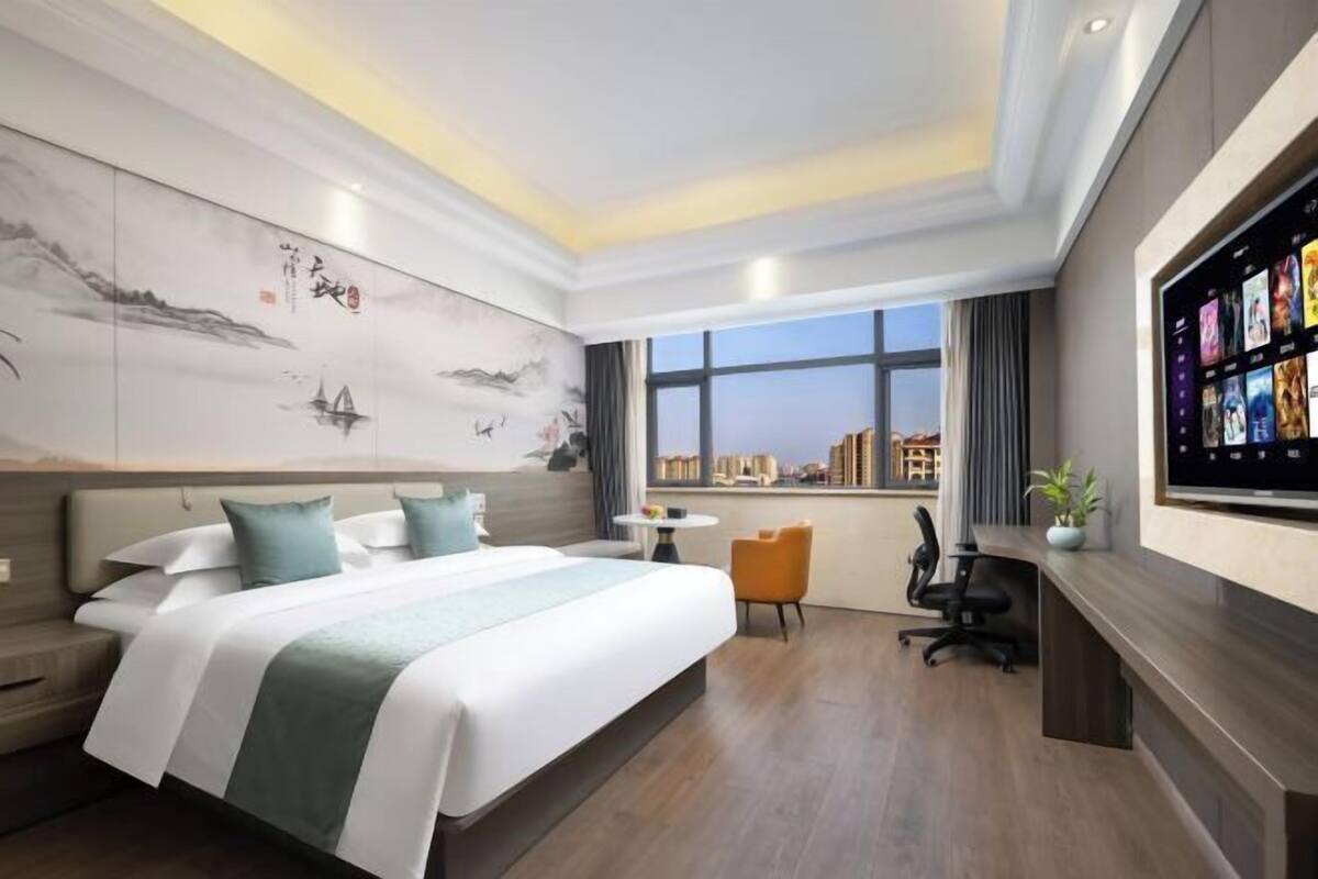 A modern hotel room with a large bed, a desk, a chair, a TV, and a city view.