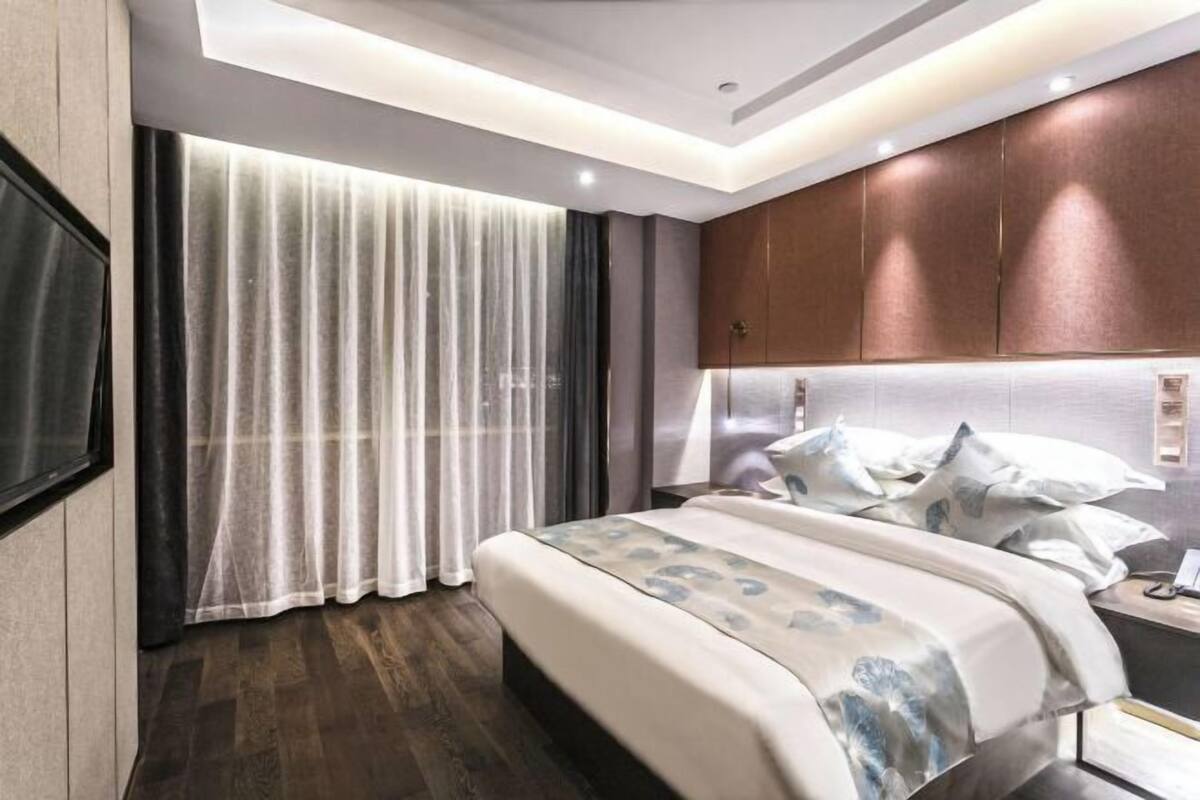 A modern hotel room with a large bed, a headboard with built-in lighting, a bedside table, and a large window with curtains.