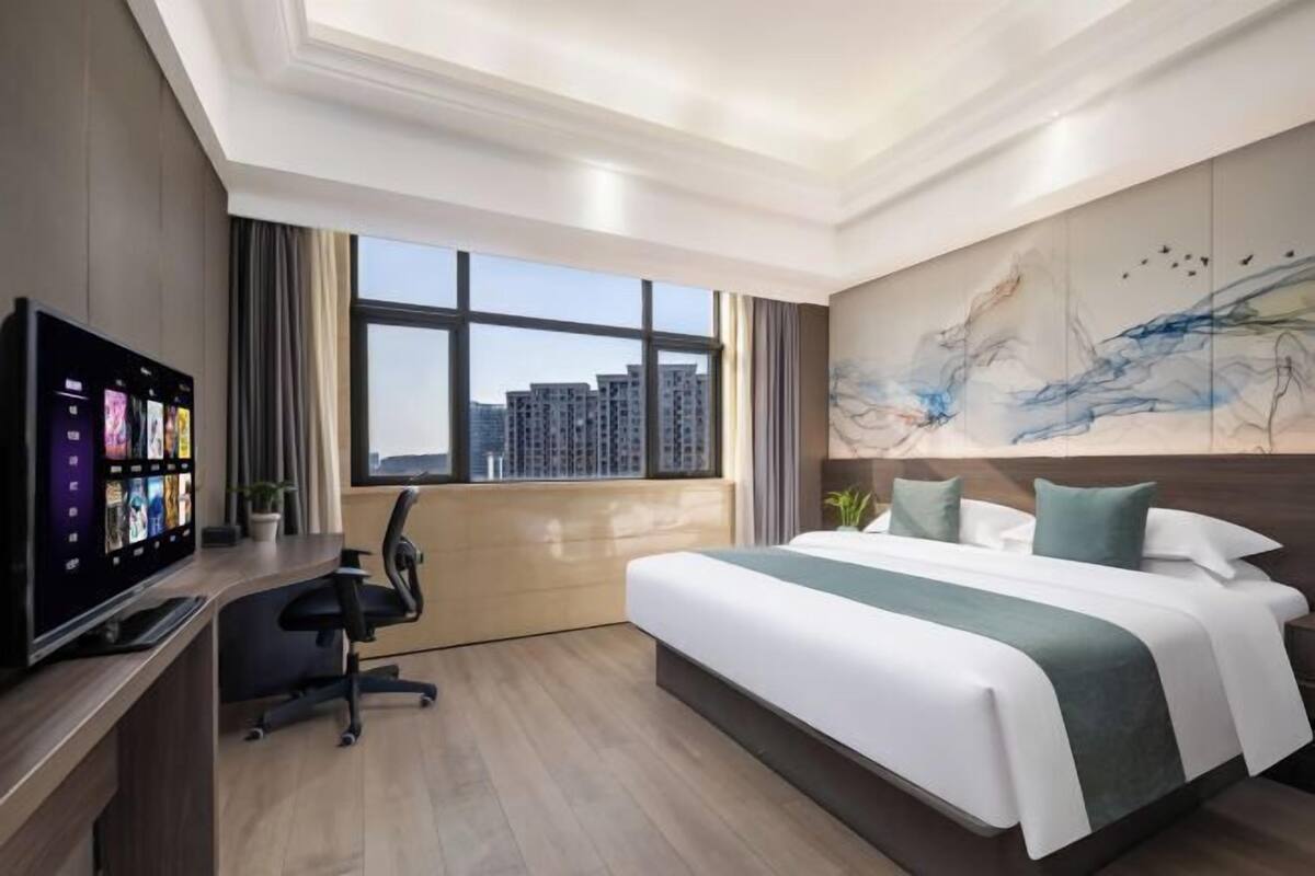 A modern hotel room with a large bed, a desk with a computer, and a city view through the window.