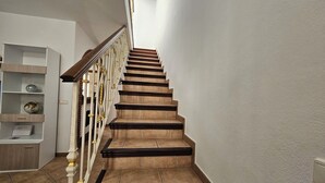 Una escalera con barandilla de madera y escalones color beige, que conduce a un rellano con pared blanca y un mueble de estantería con adornos.