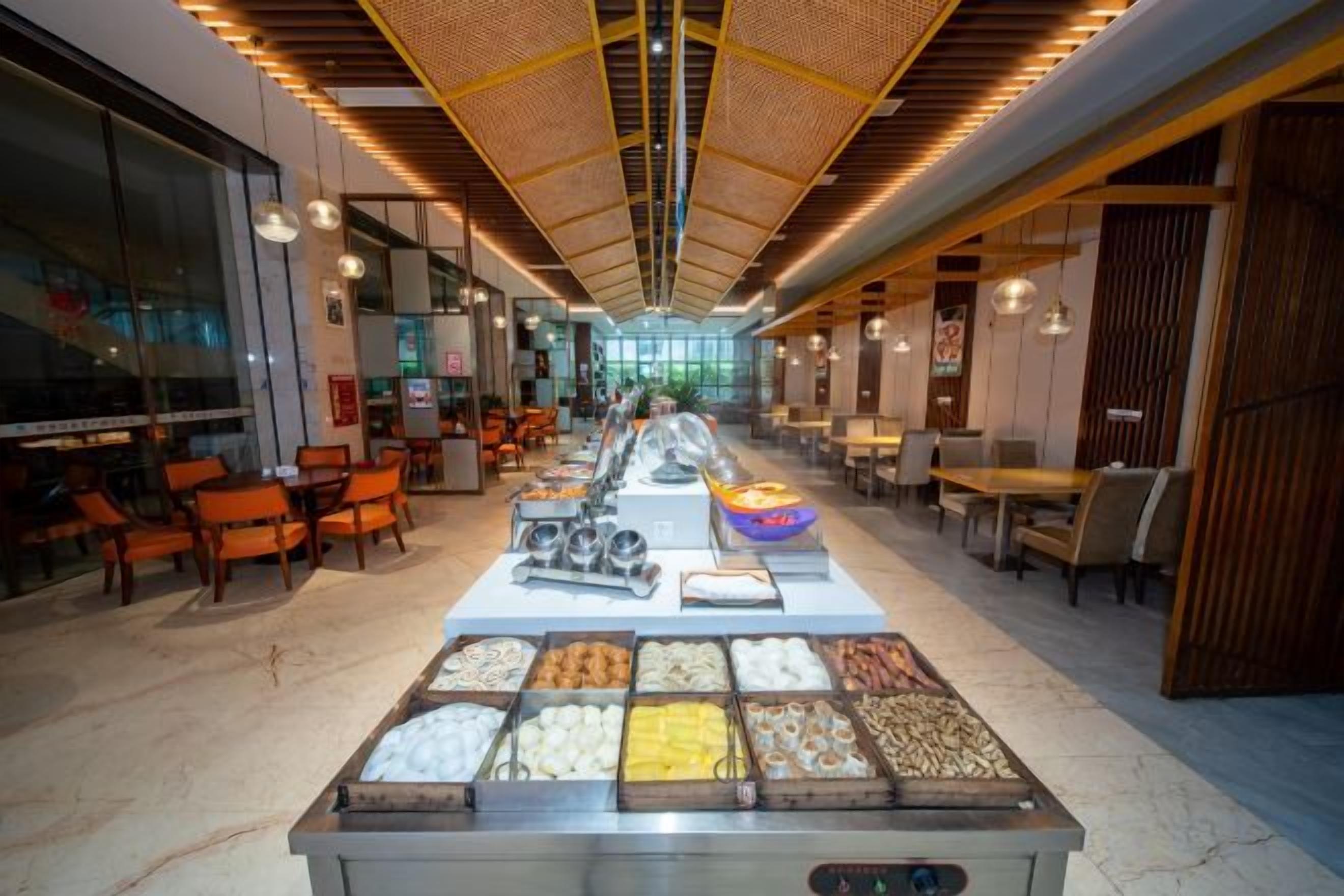 A buffet setup with various food items in the foreground, a long dining area with tables and chairs, and a modern ceiling design.